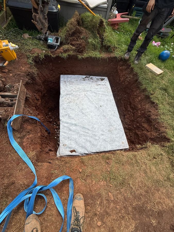 An excavation opened in a lawn and lined with geotextile membrane, ready to take stone