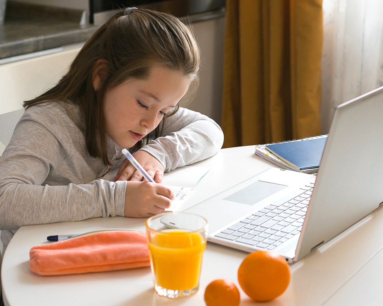 A student working at a laptop at the kitchen table, writing in a notebook