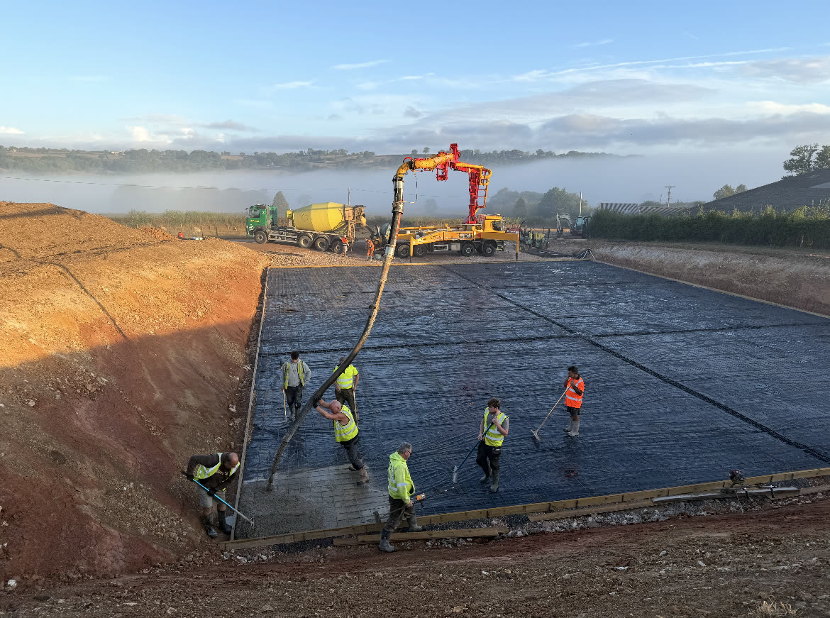 Concrete being pumped onto a large mesh-reinforced slab on a farm at dawn, with an S. Morris mixer feeding the pump and groundworkers levelling it
