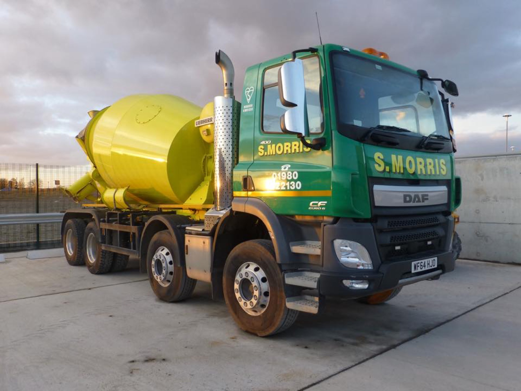 An S. Morris eight-wheel DAF mixer, green cab and yellow drum, on the concrete apron at the plant