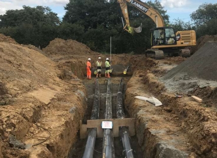 Cable ducts bedded into a trench on a cable installation, with an excavator and groundworkers behind