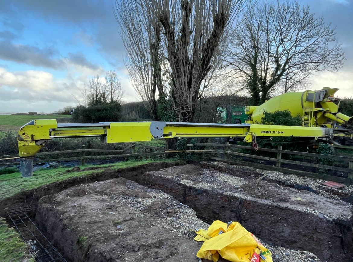 A Liebherr conveyor belt extended from an S. Morris mixer out over a fence, placing concrete into strip foundation trenches on a rural site